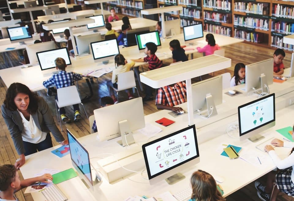 Children working on computers in a school library while a teacher helps