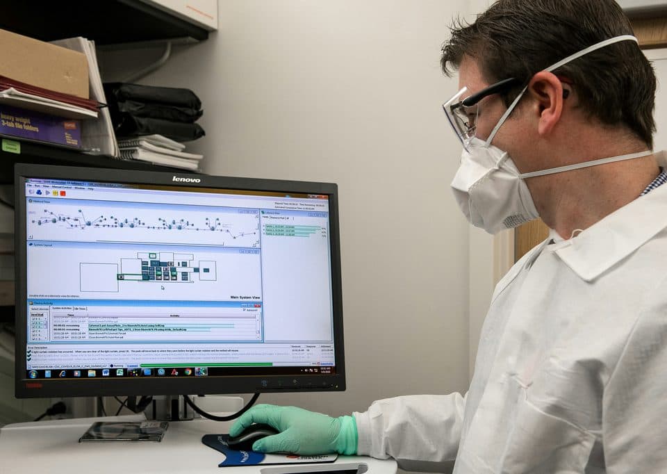 A lab technician in a mask and gloves checking results on a computer